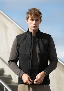 A model wearing a black unisex vest over a long-sleeve brown top, against a stairway and blue sky background.