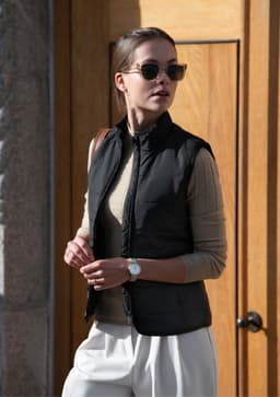 A woman wearing a black vest over a tan top, holding a bag, standing in front of a wooden door. Sunglasses complete her look.