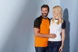 A man in an orange raglan T-shirt and a woman in a white raglan T-shirt pose together against a blue background.