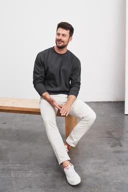 A round-neck heavy sweater in dark grey worn by a man seated on a wooden bench, paired with light trousers.