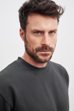 A model wearing a dark grey round-neck sweater. The sweater features a simple design and relaxed fit.