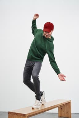 A man wearing a green hooded sweatshirt and a red cap balances on a wooden bench, dressed in grey jeans and white sneakers.