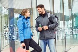 A blue softshell jacket worn by a woman and a grey softshell jacket on a man, both standing outdoors.