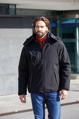 A man in a black softshell jacket with a hood, featuring a zippered pocket, walks outside.