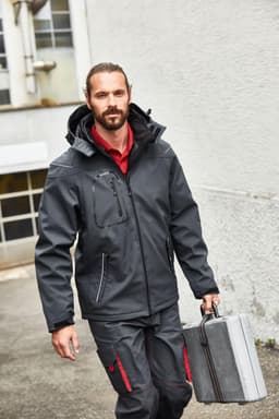 A man in a black softshell jacket carries a grey tool case, walking outdoors. The jacket features a hood and zippers.