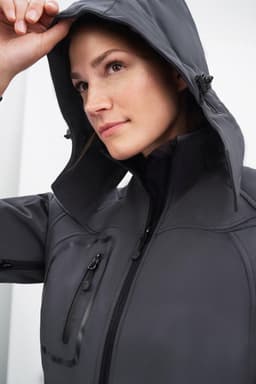 A grey ladies' softshell jacket featuring a hood, with a close-up of a woman wearing it and a logo present.
