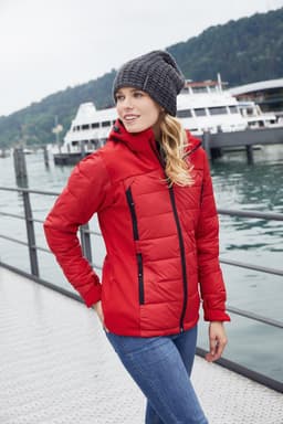 A red ladies' outdoor hybrid jacket with a hood, worn by a woman by the water, featuring a knit beanie.
