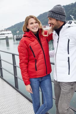 A red and a white soft shell jacket are displayed, worn by a couple against a scenic waterfront backdrop. The jackets have zippers and one features a logo.