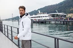 A man in a white soft shell jacket stands by a waterfront, with a boat and people in the background.