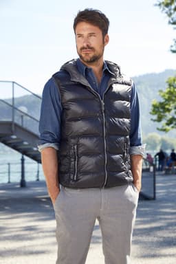 A man wearing a black down vest over a blue shirt and grey trousers, standing outdoors by a lakeside.