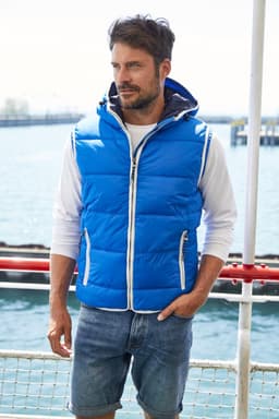 A blue padded men's vest with a hood, worn over a white long-sleeve top and paired with denim shorts.