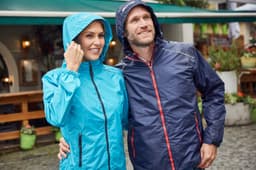 A light blue ladies rain jacket and a navy men's rain jacket, both featuring hoods, in an outdoor setting.