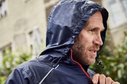 A man's navy rain jacket with a hood, featuring a zippered front and drawstring, worn in a rainy outdoor setting.