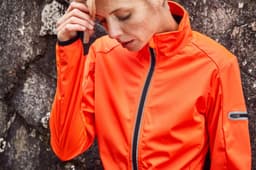 An orange ladies sports softshell jacket features a zip front and a logo, set against a textured rock background.