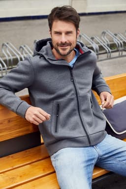 A men's hooded jacket in grey, featuring zippered pockets, worn by a man sitting on a wooden bench.