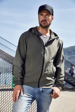 A man in a green hooded softshell jacket stands casually against an urban backdrop, wearing a cap and jeans.