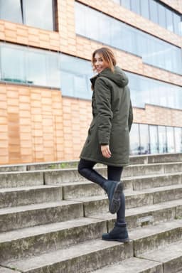 A woman wearing a green parka jacket with a hood, walking up outdoor steps. The jacket features a logo.
