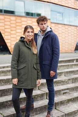 A green and a navy blue parka jacket on models standing on steps outside a modern building. Each jacket features a hood and branding.