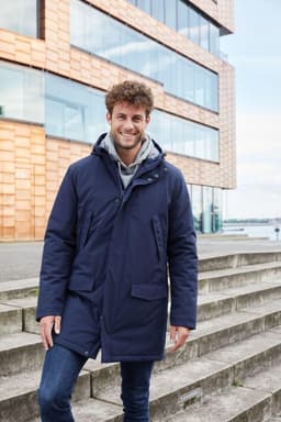 A men's business parka in navy blue, featuring a hood and pockets, worn by a smiling man on outdoor steps.
