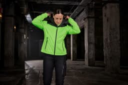 A bright green ladies all-weather jacket with a hood is worn by a person indoors, with a dark backdrop.
