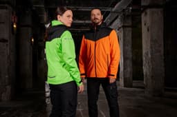 A green and black ladies rain jacket and an orange and black men's rain jacket are displayed together in an industrial setting.
