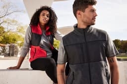 A grey and black men's fleece vest is worn by a man, while a woman in a red and black vest sits nearby.