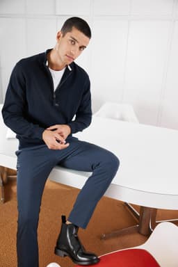 A navy half-zip jumper worn by a model, paired with navy trousers and black ankle boots, set in a modern interior.