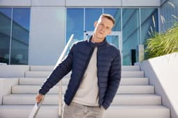 A navy padded jacket worn by a man standing on outdoor steps with modern architecture in the background.