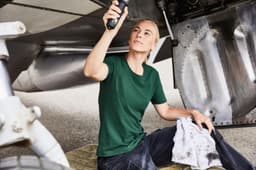 A green work t-shirt worn by a woman, seated on a mat near an aircraft's undercarriage, holding a tool.