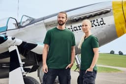 A green BIO Stretch-T shirt displayed on two models in front of a vintage aircraft. The shirt features a logo.