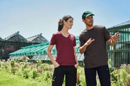 A maroon ladies' T-shirt and a brown men's T-shirt are displayed in a garden setting with greenhouses in the background.