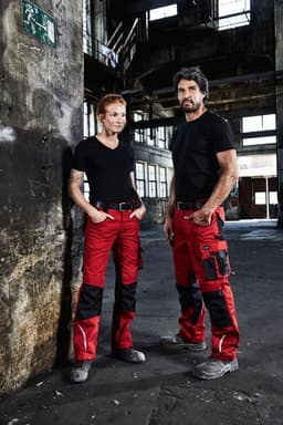A pair of individuals in black t-shirts and red work trousers with black accents, standing in an industrial setting.