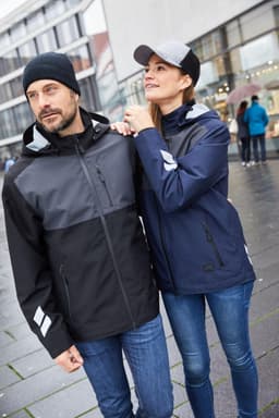 A hardshell workwear jacket in black and navy, featuring reflective details, worn by a man and a woman outdoors.