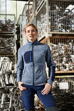 A women's polar fleece jacket in blue with a zippered front and a pocket, modelled against a backdrop of metal materials.