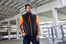 A man in a black hybrid vest with orange accents poses in a spacious indoor setting. The vest features a logo.