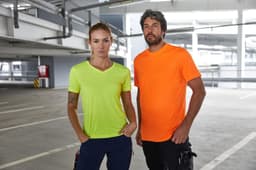 Two individuals stand together in a parking garage, wearing a neon yellow and an orange T-shirt. The shirts have a logo.