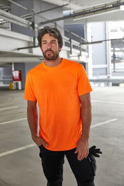 An orange work T-shirt is worn by a man standing inside a parking garage. The shirt has a logo on it.