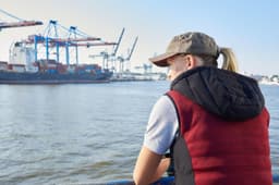 A woman in a red and black padded hybrid vest gazes at a waterfront scene with ships and cranes.