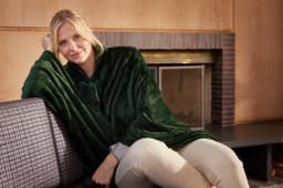 A green microfibre fleece blanket XL draped over a seated woman on a couch, with a fireplace in the background.