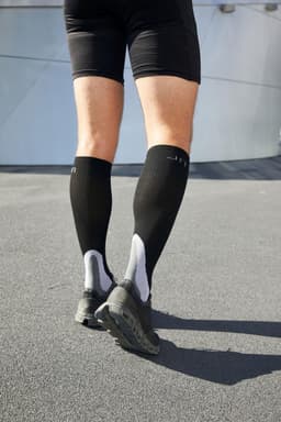 A pair of black compression socks worn with shorts and running shoes, viewed from behind on a textured surface.