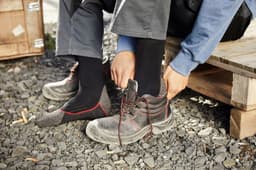 A pair of black work socks is being put on, with work boots and gravel beneath. The setting includes wooden pallets and boxes.