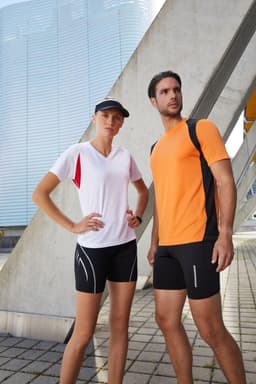 A model wears a vibrant orange men's running T-shirt paired with black shorts. The setting features modern architecture.