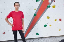 A red T-shirt is displayed on a young model against a climbing wall with colourful holds.