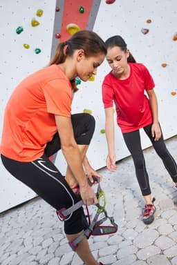 Two girls in vibrant orange and red T-shirts are preparing for a climbing activity at a rock wall.