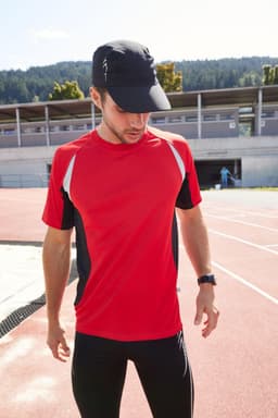 A man wearing a red and black running t-shirt with a cap, standing on a sports track.