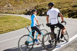 A couple in cycling attire, a blue and white jersey for the woman and a black jersey for the man, stand beside their bikes.