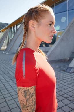 A red ladies sports T-shirt is displayed on a model with braided hair and a tattooed arm. The shirt features reflective accents.