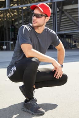 A man wearing a grey T-shirt and black leggings, complemented by a red cap and sunglasses, poses outdoors.