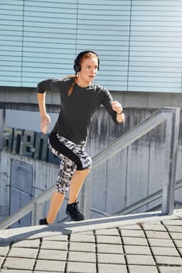 A woman wearing black and white patterned 3/4 tights and a grey top runs up stairs, wearing headphones.