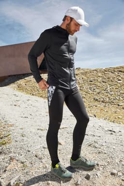 A man in a black zip hoodie and black leggings stands outdoors, wearing a white cap and sporty shoes. The background features rocky terrain.